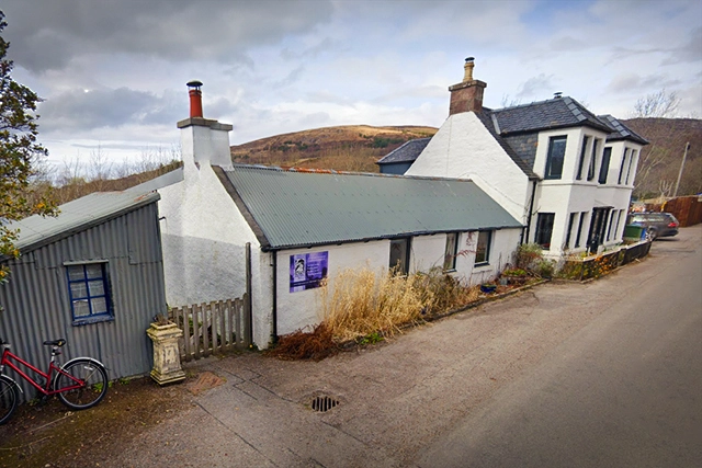 Bridgehouse Art, an independent art school in Ullapool, presents a quaint white building with an adjoining grey studio, nestled in the North-West Highlands. It sits quietly along Old Moss Road, with a red bicycle parked out front against a fence. Overgrown plants and a weathered sign indicate a place of rustic charm and creative endeavour. The rolling hills in the background offer an idyllic, inspiring Scottish setting for students seeking artistic growth. The atmosphere is one of tranquillity and potential, inviting artists to explore and express amidst nature’s beauty.