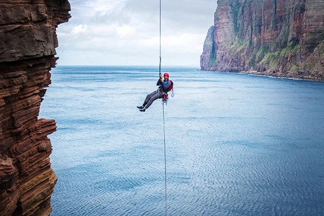 An adventurer from Hamlet Mountaineering, situated near Ullapool in Achiltibuie, is captured in a thrilling moment of abseiling down a sheer cliff face. Wearing a red helmet, a blue jacket, and secured with harnesses and ropes, the individual appears in high spirits despite the challenging activity. The surrounding landscape boasts dramatic vertical cliffs that rise majestically from the deep blue sea. The image conveys a sense of excitement and the vast scale of nature, encapsulating the exhilarating experiences offered by the company, from rock climbing to hiking and more in the rugged Scottish terrain.