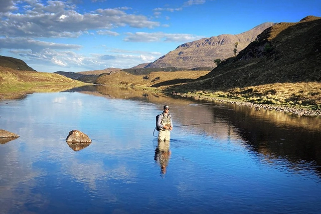 A serene image of a fly fisherman, likely James Curly, engaged in trout and salmon fishing in the waters around Assynt, near Ullapool. He stands mid-river, surrounded by calm waters reflecting the blue sky. The fisherman is dressed in appropriate outdoor gear, focused on his fishing technique. In the background, majestic hills and mountains under the bright sky create a stunning backdrop for this peaceful activity. The environment conveys the tranquillity and beauty of fishing in the Scottish Highlands, ideal for those seeking lessons or a day out on the water.