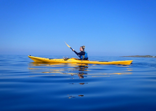 In this serene image, a person engages in a kayaking adventure on the clear blue waters of the northwest coast of the Scottish Highlands, provided by 'Summer Isles Sea Kayaking' in Ullapool. They're in a vibrant yellow kayak, holding a double-bladed paddle mid-stroke. The kayaker, wearing a blue life jacket, appears relaxed and focused, enjoying the calm sea under a clear sky. The water is so still that it mirrors the kayak and the paddler, creating a sense of peaceful symmetry, perfect for family days out and learning about kayaking.