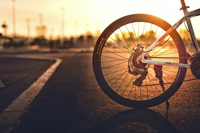 This image captures the essence of a cycling adventure provided by a bike hire company in Ullapool, focusing on the rear wheel of a bicycle. The setting sun casts a warm golden glow on the scene, highlighting the details of the wheel and its intricate spokes. The bike is poised on smooth asphalt, with the blurred outline of a parking lot and distant street lamps in the background. The ambiance suggests an inviting end to a day filled with exploration and the simple joys of bike riding in the picturesque surroundings of Ullapool.