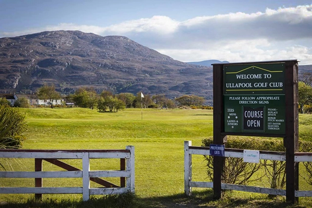 The entrance to Ullapool Golf Club, a 9-hole course situated along the shore of Loch Broom. The welcoming signboard at the foreground reads 'Welcome to Ullapool Golf Club' with additional notices including directions and course status. The well-maintained green fairways are set against the backdrop of rolling hills and mountains under a clear blue sky with scattered clouds. A traditional white fence opens to invite players onto the course, reflecting a tranquil setting for golf enthusiasts to enjoy a round in the scenic landscape of Ullapool.