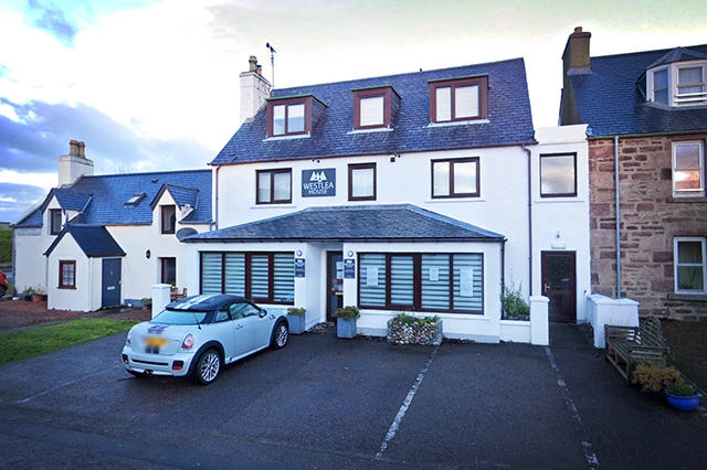 Westlea House, a boutique Bed & Breakfast in Ullapool, is presented in this image as a two-story white building with a contrast of dark roofing and window frames. The B&B boasts a pristine and inviting exterior with a sign that features its name prominently. In front of the building is a well-maintained parking area with a light blue convertible car, adding to the charm of the guesthouse. The surrounding environment is peaceful, with a backdrop of gentle hills, indicating its proximity to the scenic harbour front. This image conveys a sense of the establishment's elegance, comfort, and welcoming hospitality that awaits its guests.