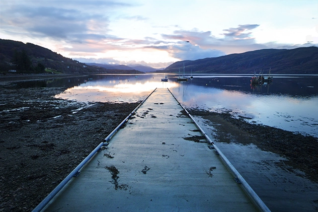The image showcases a tranquil scene at a small pier or jetty in Ullapool, extending into Loch Broom, known as the base for WhatSUP Ullapool paddle boarding activities. The jetty runs straight into the calm reflective waters, which mirror the pastel hues of the dawn or dusk sky. Mountains gracefully rise in the distance, framing the serene loch. A few boats are moored in the still water, adding to the picturesque setting. Bird tracks are visible on the jetty's surface, hinting at the wildlife that frequents this peaceful spot.