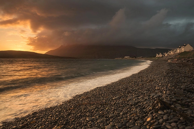 A stunning sunset view at Ardmair Beach, located just outside Ullapool, offering a serene and inviting addition to the list of things to do in Ullapool. The beach features a shore lined with smooth pebbles, leading to gentle waves of the sea. The dramatic sky, with clouds lit by the golden light of the setting sun, casts a warm glow over the landscape, while the distant mountains create a striking silhouette against the fiery sky. This tranquil scene is a beautiful representation of Scotland's natural coastal beauty.
