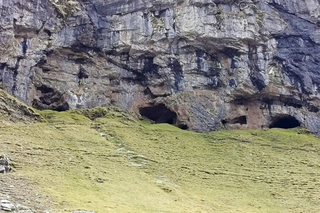 A photo showcasing the rugged entrance of the historic Inchnadamph Bone Caves near Ullapool, nestled within the towering limestone cliffs. The caves, visible as dark openings on the mountainside, are set against a backdrop of lush green slopes. These ancient caverns, steeped in archaeological significance, are a silent testament to Scotland's prehistoric past, offering modern explorers a portal into the lives of the animals and humans that once sought refuge in their depths.