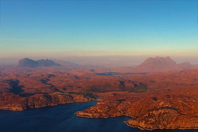 A captivating aerial view of the Assynt and Coigach landscapes near Ullapool, with the prominent peaks of Suilven and Cul Mor rising majestically in the distance. The terrain is a tapestry of rich, russet-toned moorland interspersed with glistening lochs. This vista captures the serene beauty of the Scottish Highlands during what appears to be the golden hour, with the sun casting a warm glow over the rugged scenery.