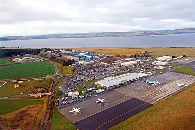 Aerial view of Inverness Airport, the nearest airport to Ullapool, located in the scenic Highlands of Scotland. The image shows the airport's expansive layout with its main runway and several parked aircraft, including both commercial jets and smaller private planes. The surrounding area features a mixture of parking lots, industrial buildings, and green fields, with the Moray Firth's wide expanse visible in the background. This view highlights the airport’s role as a key hub for travellers heading to Ullapool and the North West Highlands, providing a gateway to this picturesque region.