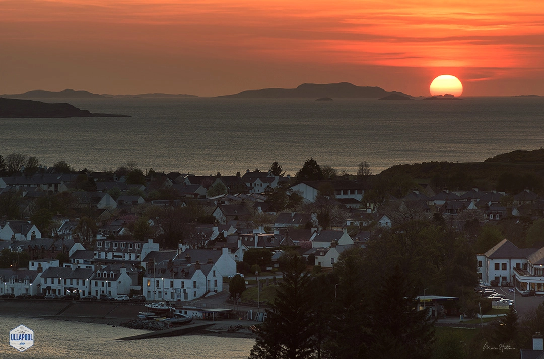The sun sets in a spectacular display of warm hues, casting a radiant glow over the village of Ullapool. In the foreground, the quaint houses and winding streets of the village begin to light up as evening approaches. Photographed by Marc Hilton from Ullapool. The shimmering waters of the sea gently lead the eye towards the silhouetted Summer Isles on the horizon, enhancing the peaceful end-of-day atmosphere. This moment captures the serene transition from day to night in the Scottish Highlands.