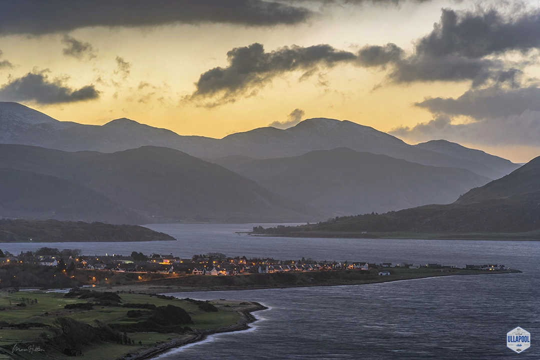 Dawn's first light gently illuminates Ullapool, as the quaint Scottish village awakens along the shimmering shores of Loch Broom. Silhouettes of rolling hills ascend into the horizon, creating a layered tapestry of natural contours in the Scottish Highlands. The glowing lights of homes and streets softly punctuate the predawn tranquillity, offering a serene moment before the sun graces the sky above the mountains. Photographed by Marc Hilton from Ullapool.