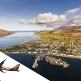 This composite image features an aerial view of Ullapool and Loch Broom in the Scottish Highlands, with a digitally superimposed airplane flying over a stylized white stripe that bisects the photo diagonally. Ullapool is depicted with its characteristic layout along the shoreline, showcasing dense residential areas, the harbour with docked ships, and the winding roads that meander through the village. The clear skies and calm waters reflect the tranquil atmosphere of the region. In the backdrop, rolling hills and a patchwork of greenery highlight the natural beauty of the Highlands, while the airplane symbolizes the access to this remote location, relating to the blog post's topic of the nearest airport to Ullapool.