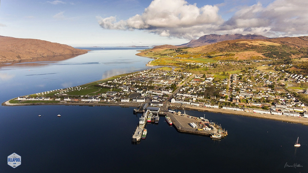 Aerial view of Ullapool, a charming village along the shores of Loch Broom in the Scottish Highlands, featuring the tranquil waters with boats, the picturesque Summer Isles in the distance, and the majestic Ben Mor Coigach mountain looming in the backdrop. Photographed by Marc Hilton of Ullapool. The dense arrangement of houses and the bustling harbour depict a lively community amid the serene natural beauty, encapsulating the essence of Ullapool's landscape and heritage.