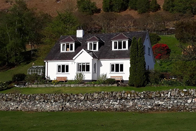 The front exterior of Clachan Farmhouse B&B, a charming two-story white house with a dark grey tiled roof, located in a picturesque rural setting. The farmhouse features dormer windows on the upper floor and a sun porch on the ground floor. The property is surrounded by lush green lawns, a stone wall, and mature trees, with a backdrop of hills and a vibrant red flowering shrub. Two wooden benches are placed on the front lawn, offering a peaceful spot to relax and enjoy the tranquil surroundings. Located 7 miles south of Ullapool.