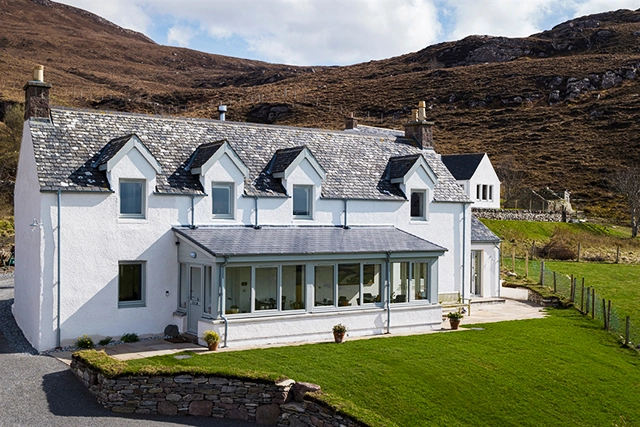 The front exterior of Croft Cottage, a charming two-story white house with a grey tiled roof, situated in a picturesque landscape at Ardmair, a few miles north of Ullapool. The cottage features several dormer windows on the upper floor, a large sun lounge with panoramic windows on the ground floor, and a small patio area with potted plants. The property is surrounded by lush green lawns, stone walls, and rugged hills in the background, creating a serene and inviting atmosphere. The clear sky and sunlight enhance the beauty of the setting, making it an ideal retreat for visitors.