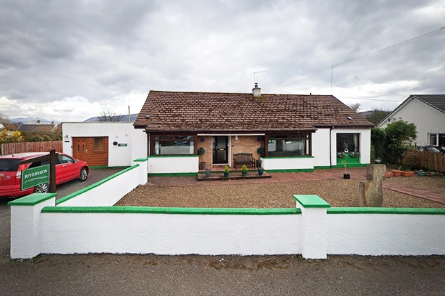 Front view of the Island View House Apartment in Ullapool, showcasing a charming single-story building with a red-tiled roof and white exterior walls accented by vibrant green trim. The property features a neatly landscaped front yard, bordered by a distinctive white and green fence, enhancing its inviting suburban appearance. The driveway hosts a red company car, indicating active hospitality services. The serene backdrop of mountainous landscapes and cloudy skies complements the peaceful residential setting, making it an attractive base for exploring the local cultural and natural attractions.
